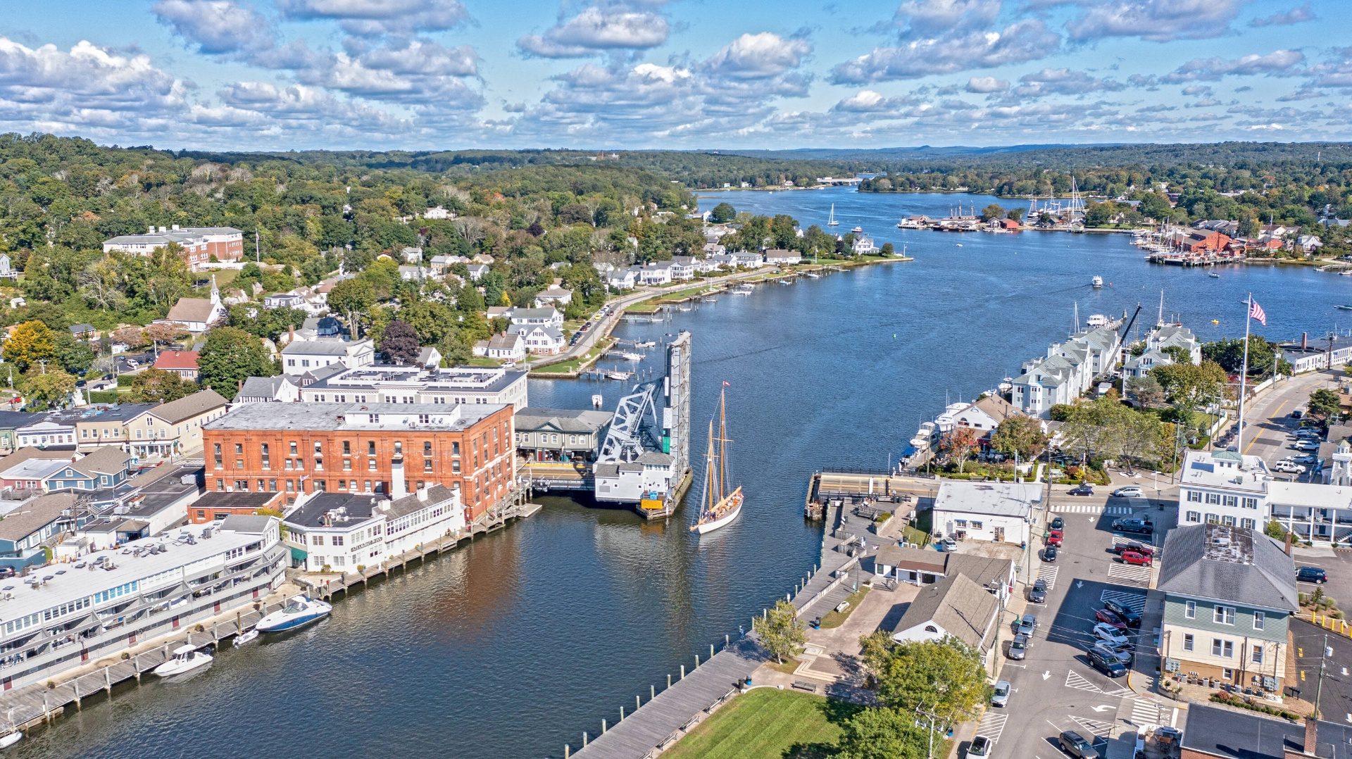 Aerial view down the Mystic River with a tall-ship schooner sailing under the open 1922 bascule drawbridge, downtown Mystic waterfront on the left and Mystic Seaport tall ships in the distance