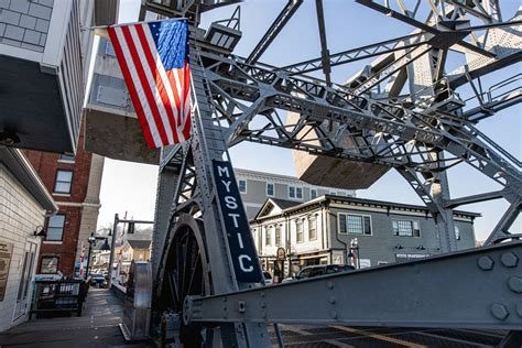 American flag waving in front of the dark iron lattice of the Mystic bascule bridge against a blue sky